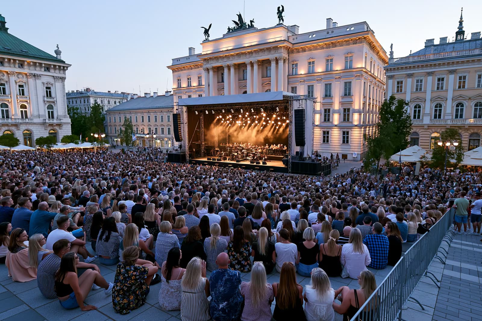 Al anochecer, una multitud se congrega en una histórica plaza de Berlín, donde un escenario al aire libre para orquesta ilumina las imponentes fachadas de los edificios.