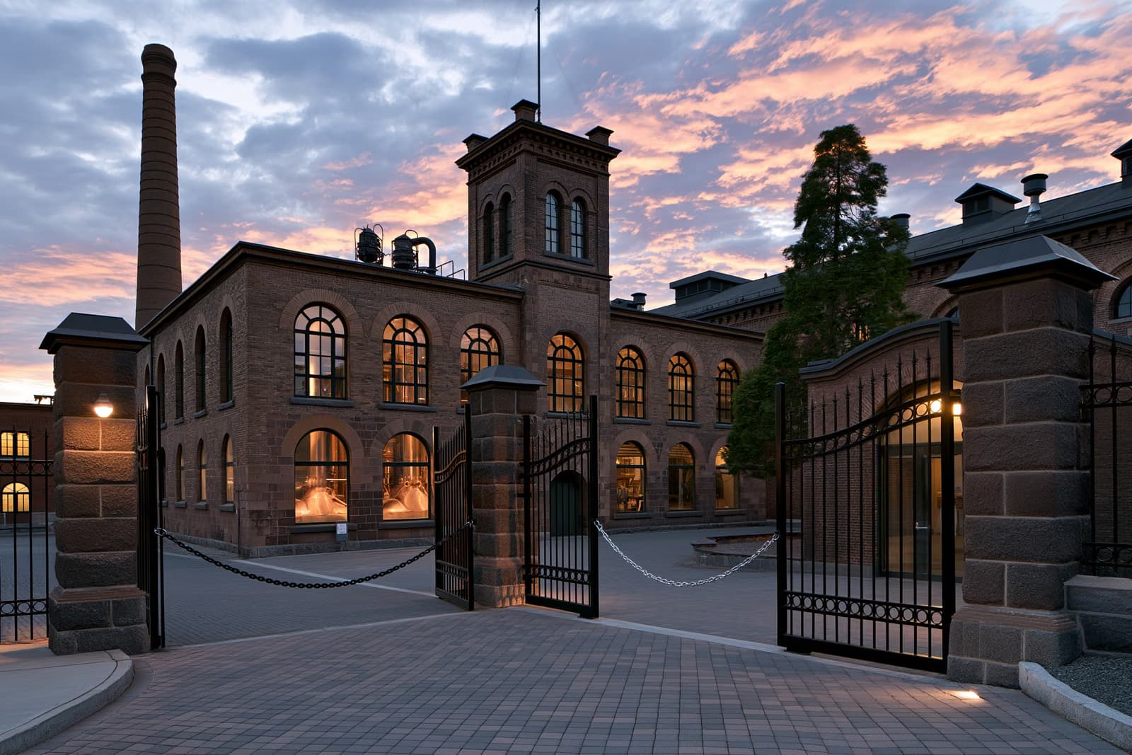 Un edificio histórico de la antigua cervecería, con calderas de cobre visibles a través de ventanas arqueadas, se alza tras unas puertas encadenadas al atardecer, marcando el final de la cervecería Eichbaum.