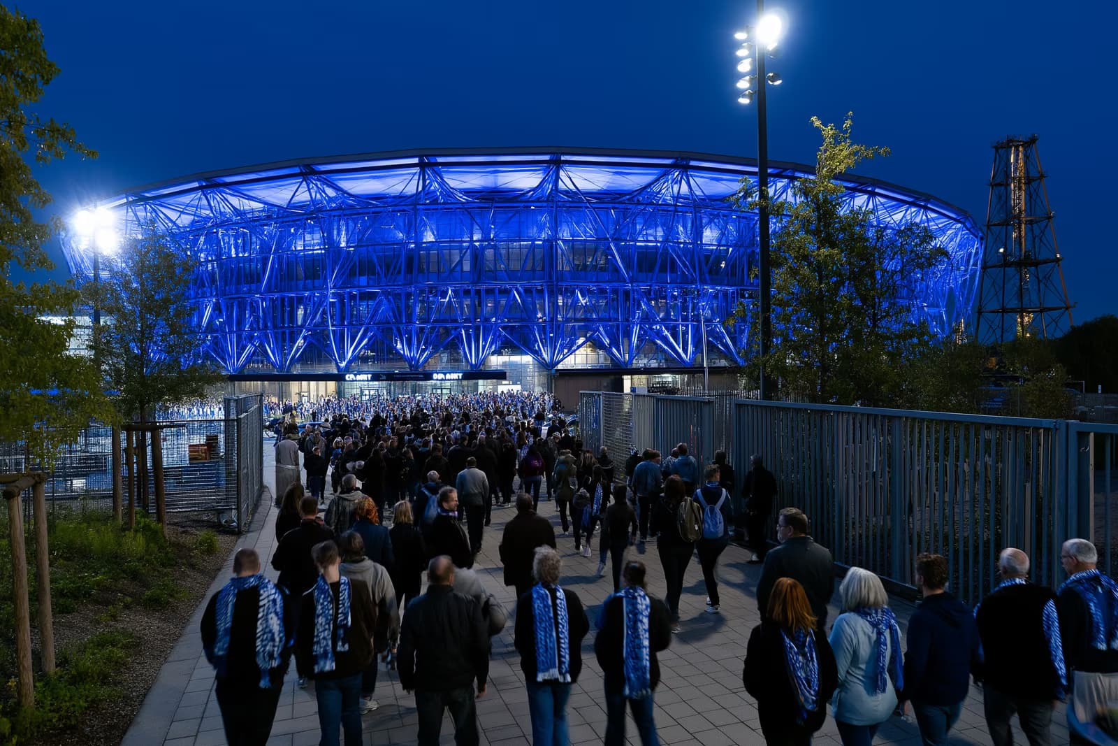 Un estadio de fútbol en la región del Ruhr brilla con un azul intenso por la noche mientras los aficionados con bufandas se agolpan hacia las puertas tras el traslado del Gosens Schalke.