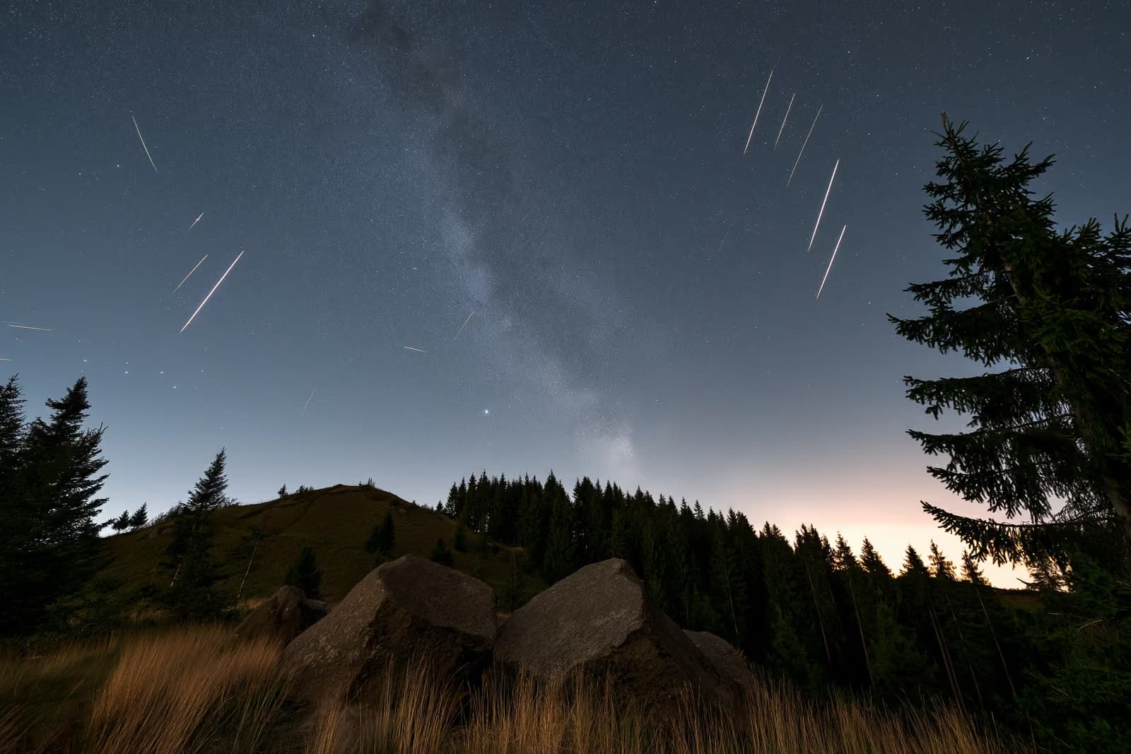Un cielo nocturno sobre un oscuro paisaje rural alemán con estelas de meteoros y una tenue banda de estrellas en lo alto.
