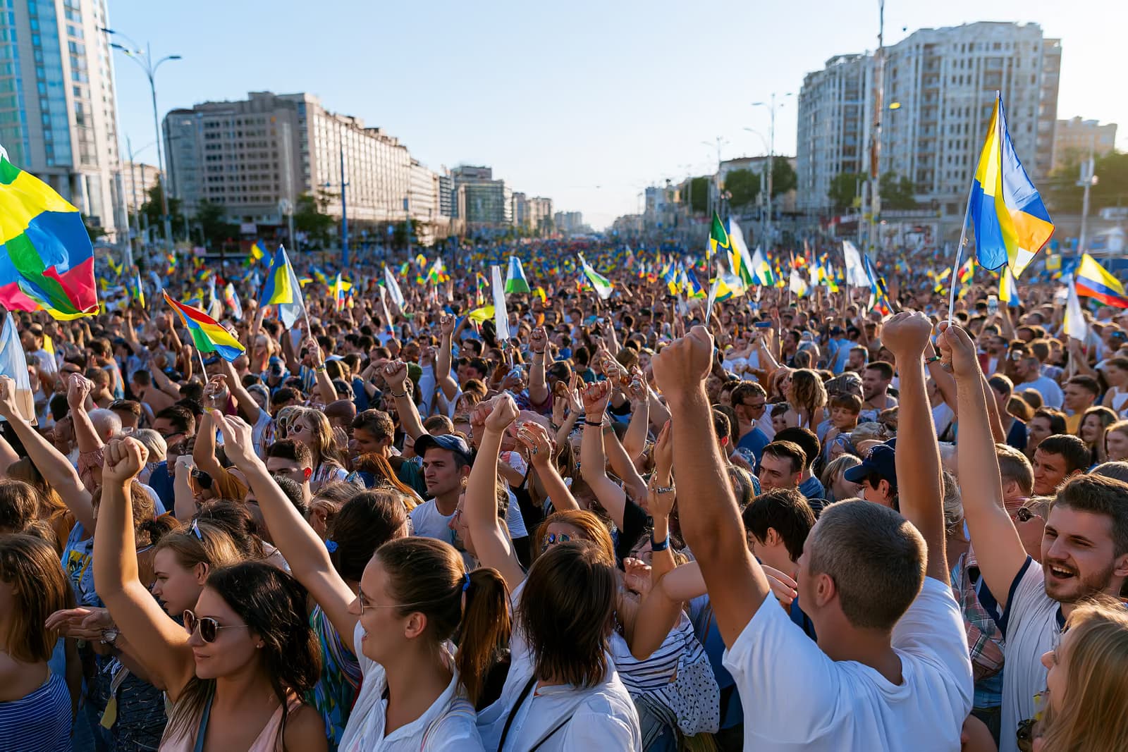 Una enorme multitud llena una calle de la ciudad bajo un brillante cielo de verano, ondeando coloridas banderas.