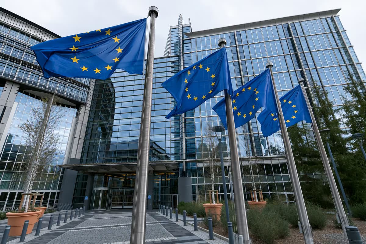 Banderas de la Unión Europea ondeando en mástiles frente a un moderno edificio de cristal.